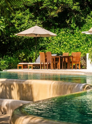 Área de piscina com cadeiras e mesa de madeira, cercada por vegetação exuberante.