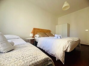 Quarto de hotel com duas camas, uma mesa de cabeceira iluminada por um abajur, um guarda-roupa e uma luminária pendente.