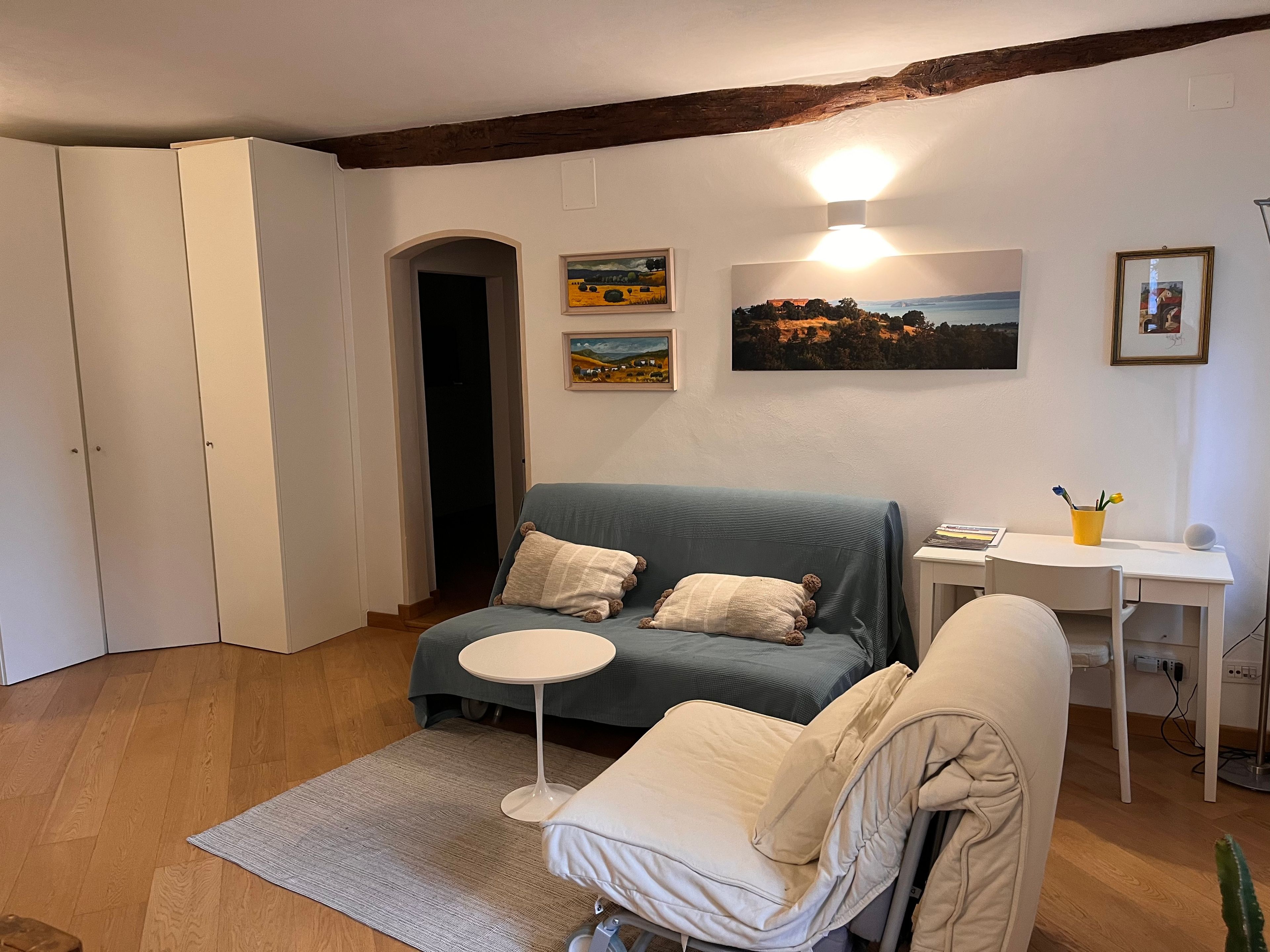 Ein gemütliches Wohnzimmer mit einem blauen Sofa, einem kleinen runden Tisch und einem weißen Sessel. Es gibt einen Holzboden, eine weiße Tür und ein Bild an der Wand.