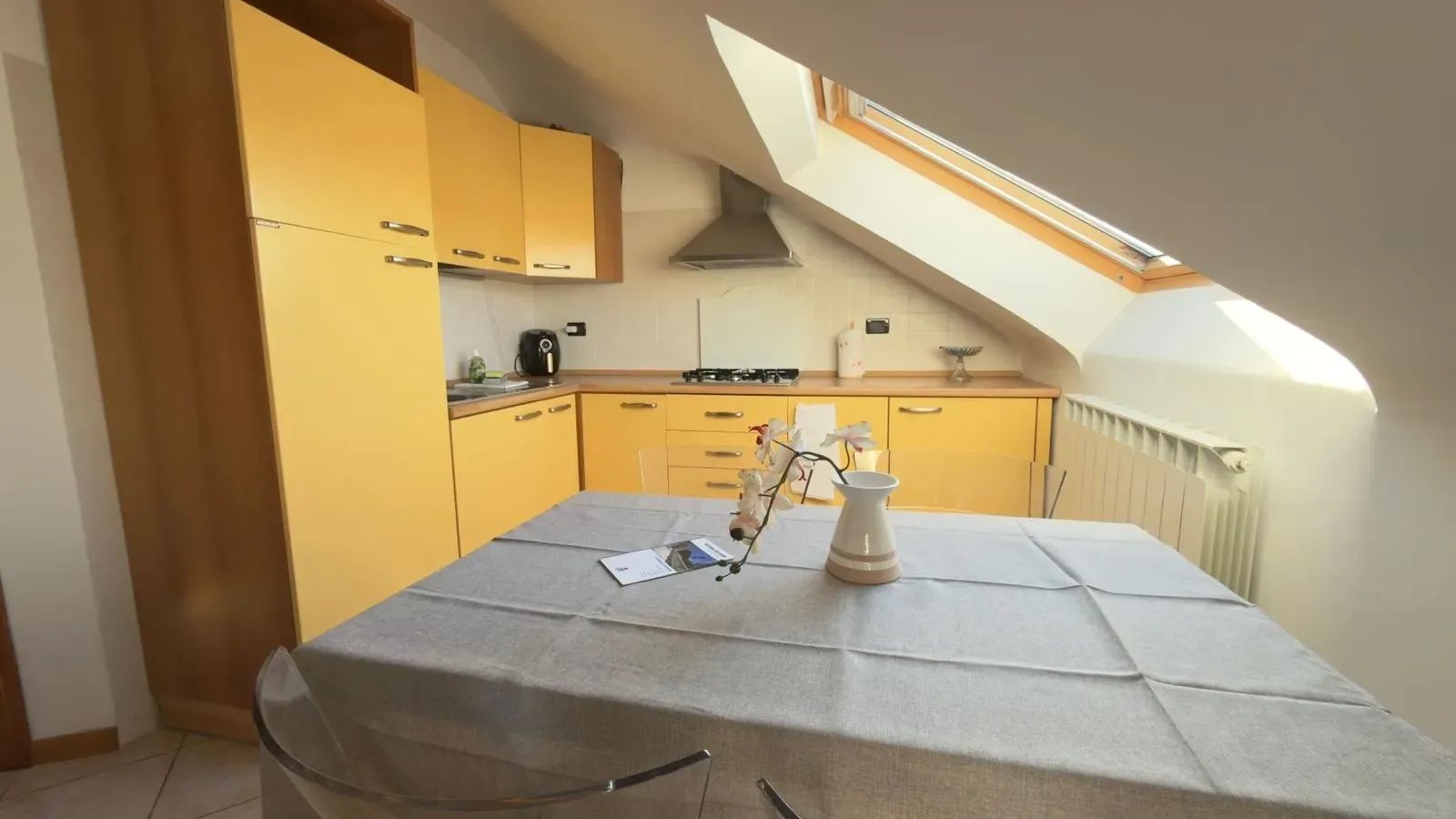 Een kleine keuken met gele kastjes, een eettafel met een grijze tafelkleed en een dakraam erboven.