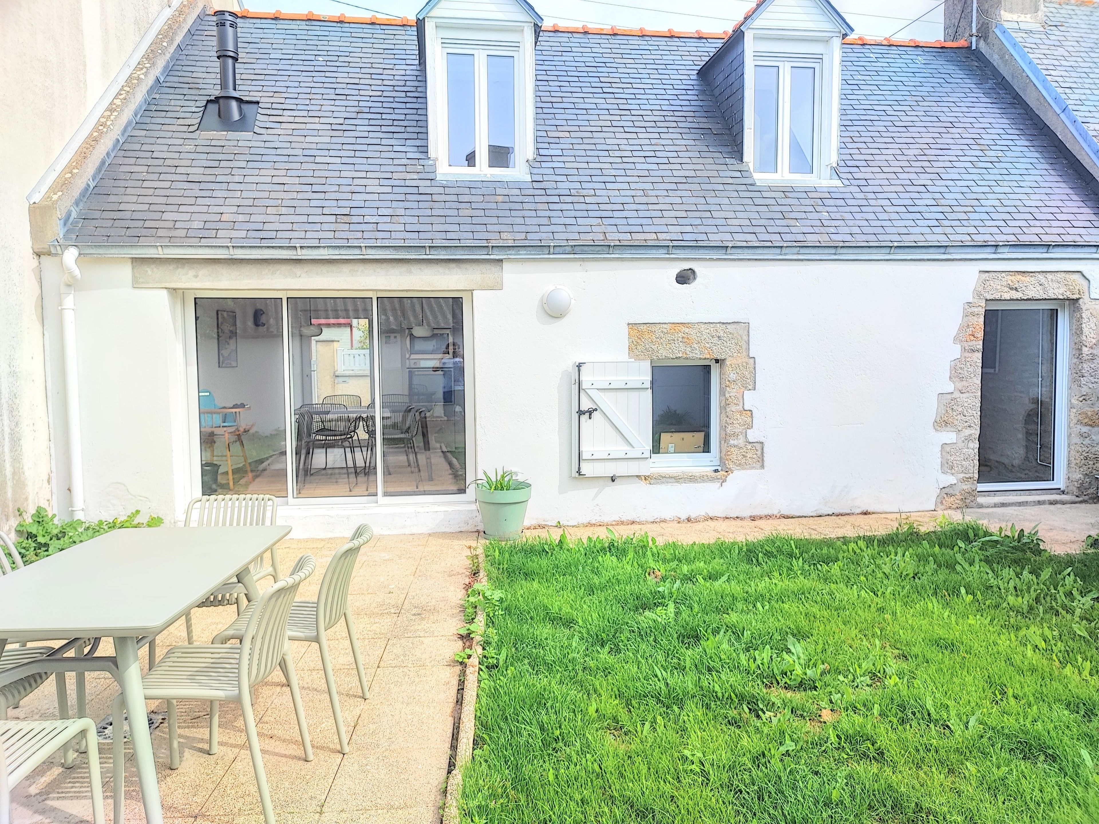 Une maison avec un toit en tuiles, une porte-fenêtre coulissante, un petit jardin et un coin repas avec des chaises et une table.