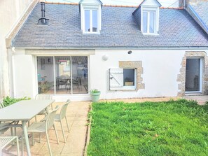 Une maison avec un toit en tuiles, une porte-fenêtre coulissante, un petit jardin et un coin repas avec des chaises et une table.