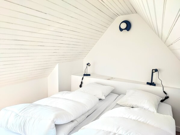 Une chambre avec un plafond en pente, deux lits avec du linge de lit blanc et deux lampes de lecture fixées au mur.