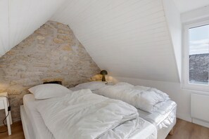 A bedroom with a sloped ceiling, a stone wall, a bed with white bedding, a bedside table with a lamp, and a window with a view of a roof.