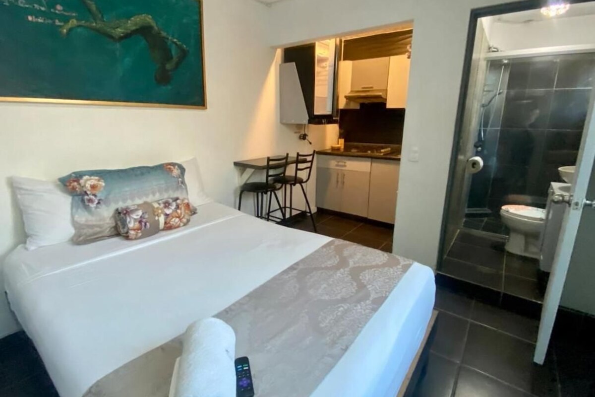A hotel room with a bed, a desk, and a view of the kitchen and bathroom.