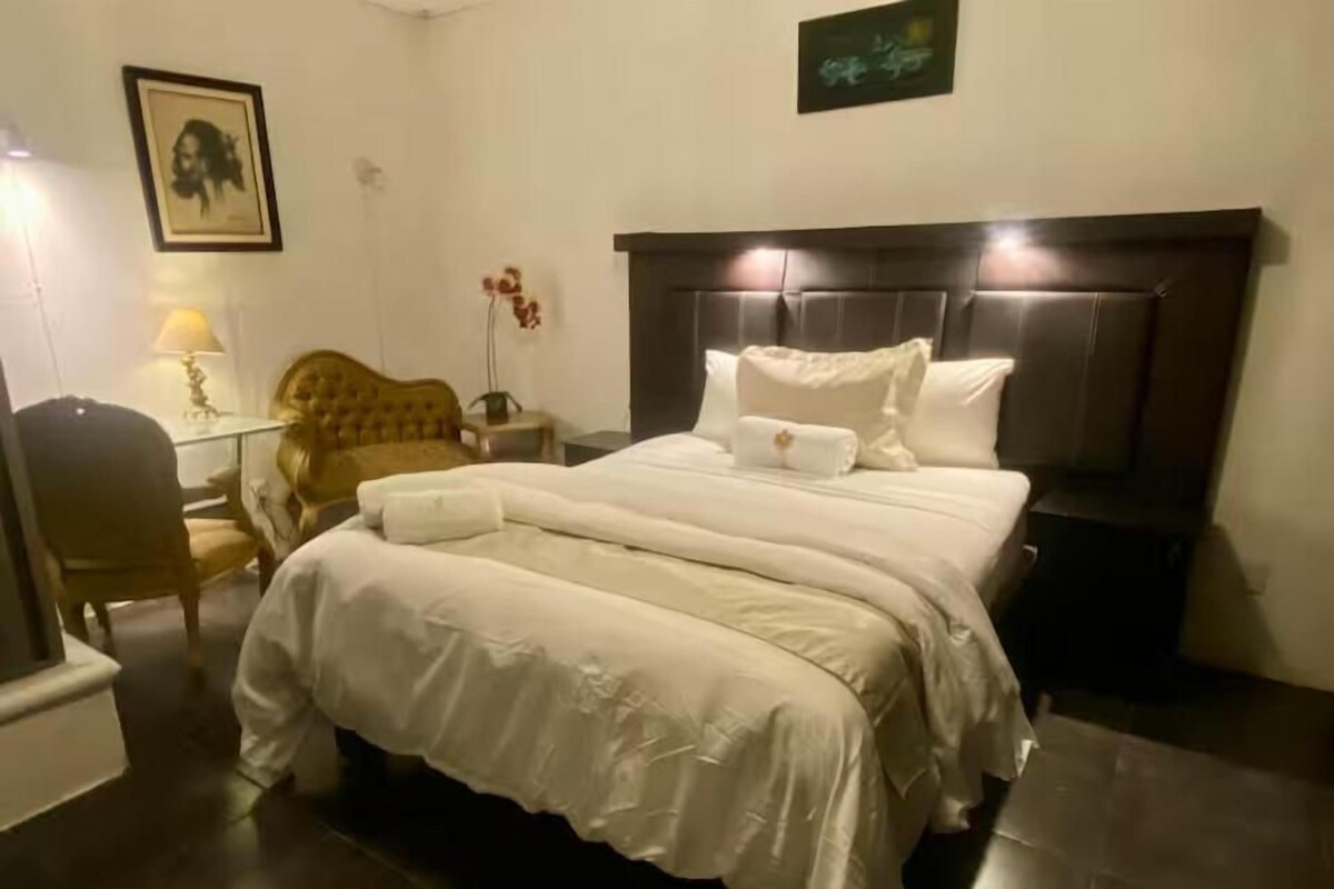 A hotel room with a large bed, a nightstand, a lamp, a chair, and a framed picture on the wall.