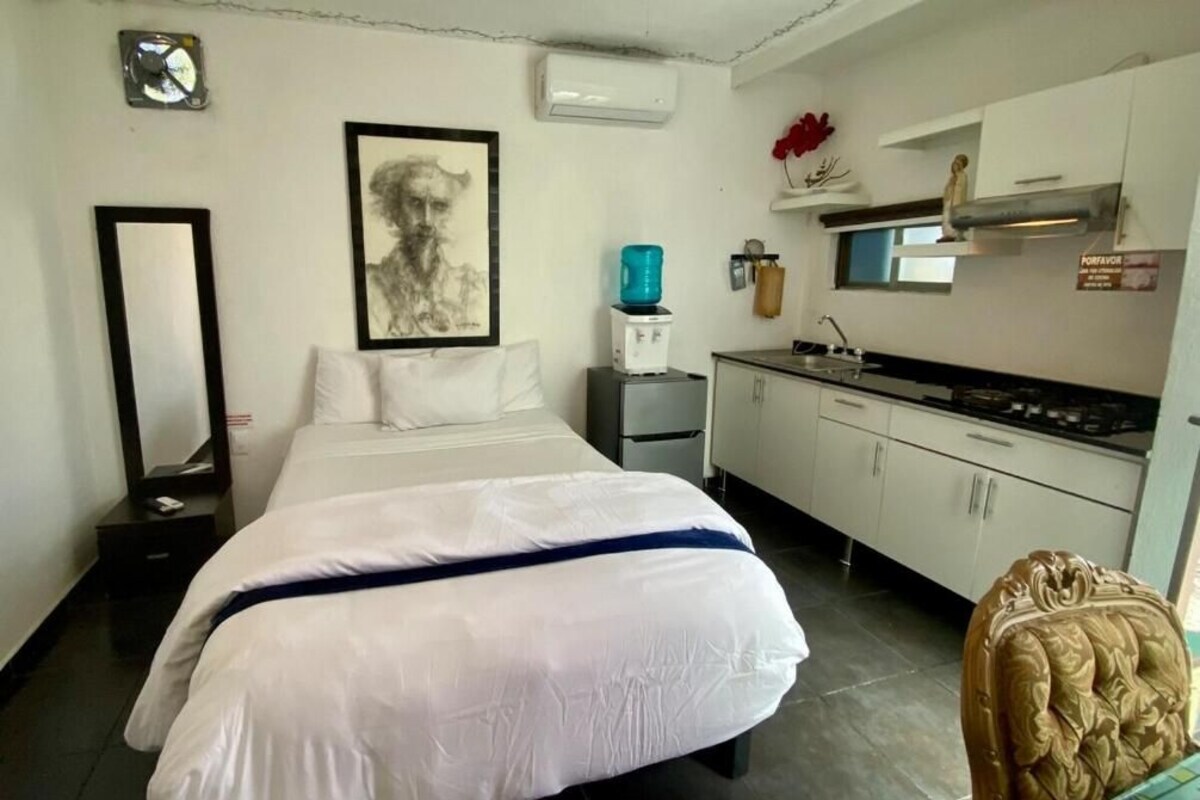 A hotel room with a bed, a small kitchenette, a mirror, and a framed artwork on the wall.