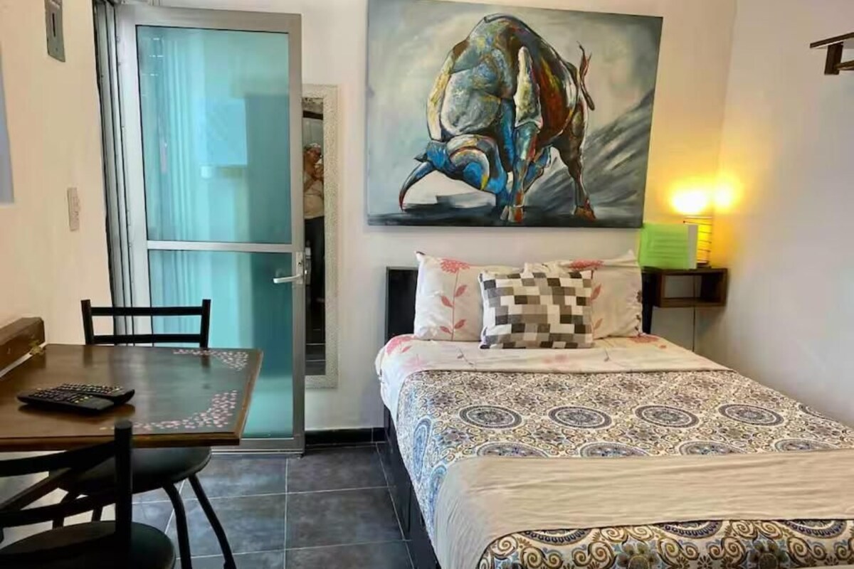 A hotel room with a bed, a desk with a chair, a painting on the wall, and a nightstand with a lamp.