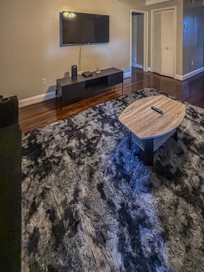 Una sala de estar con un televisor de pantalla plana montado en la pared, un mueble de entretenimiento negro, una mesa de centro redonda y una alfombra mullida.