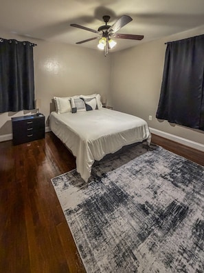 Un dormitorio con cama, ventilador de techo, mesita de noche, cómoda y alfombra estampada.