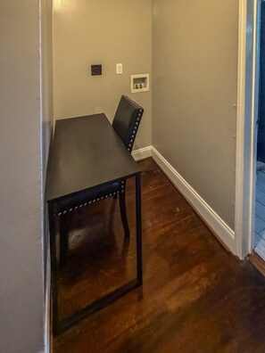 Una habitación con una mesa y silla de madera oscura, un enchufe en la pared blanca y una puerta que da a otra habitación.