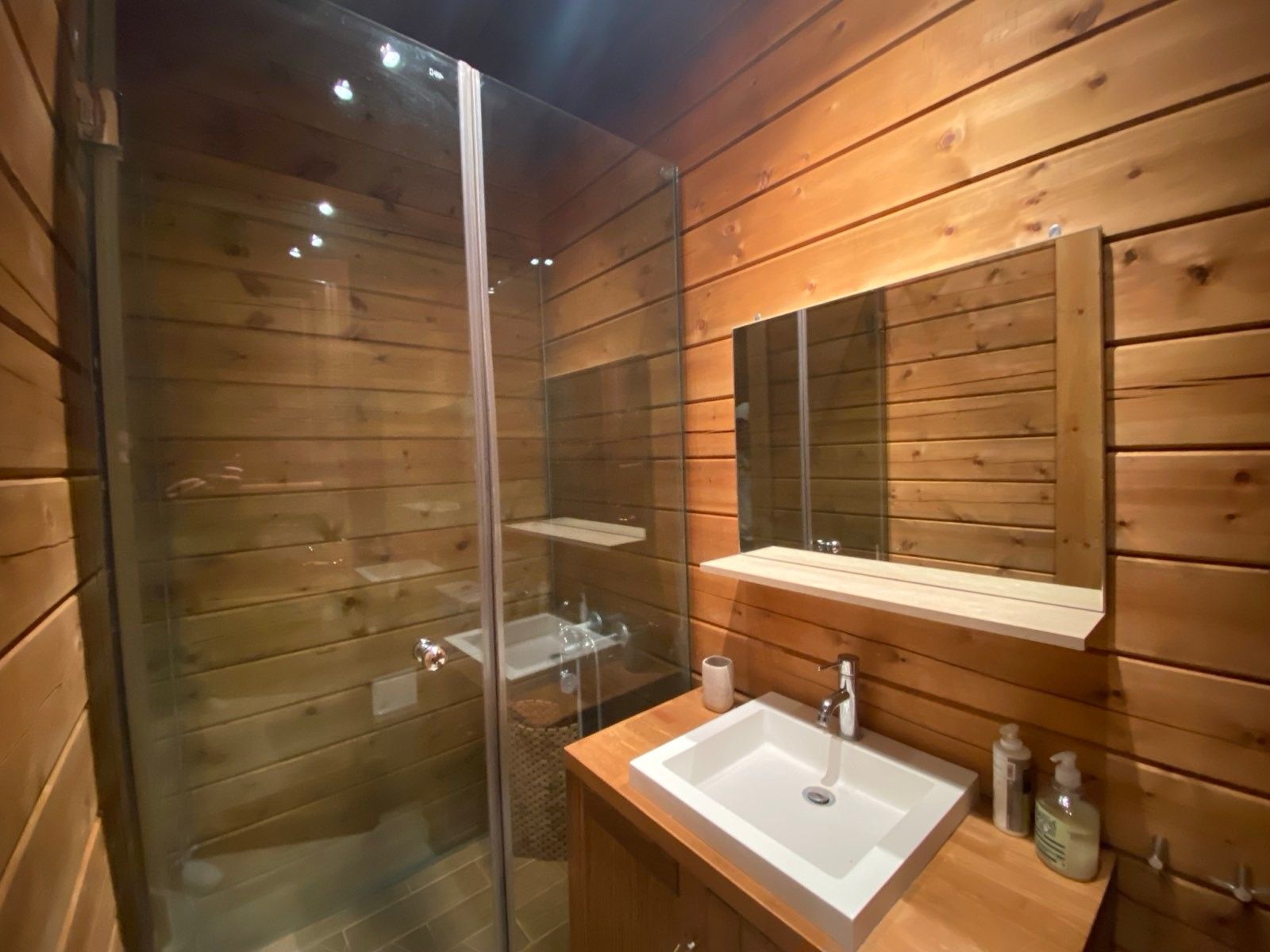 A wooden cabin bathroom with a glass shower enclosure, a sink, and a mirror.