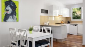 Una cocina moderna con armarios blancos, una zona de comedor con sillas blancas, una pequeña isla y un cuadro de un perro en la pared.