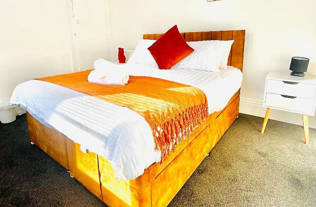 Una cama con cabecera de madera, ropa de cama blanca y un cojín rojo. Hay una mesita de noche blanca con una lámpara y un pequeño florero.