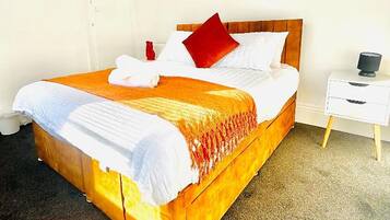 Una cama con cabecera de madera, ropa de cama blanca y un cojín rojo. Hay una mesita de noche blanca con una lámpara y un pequeño florero.