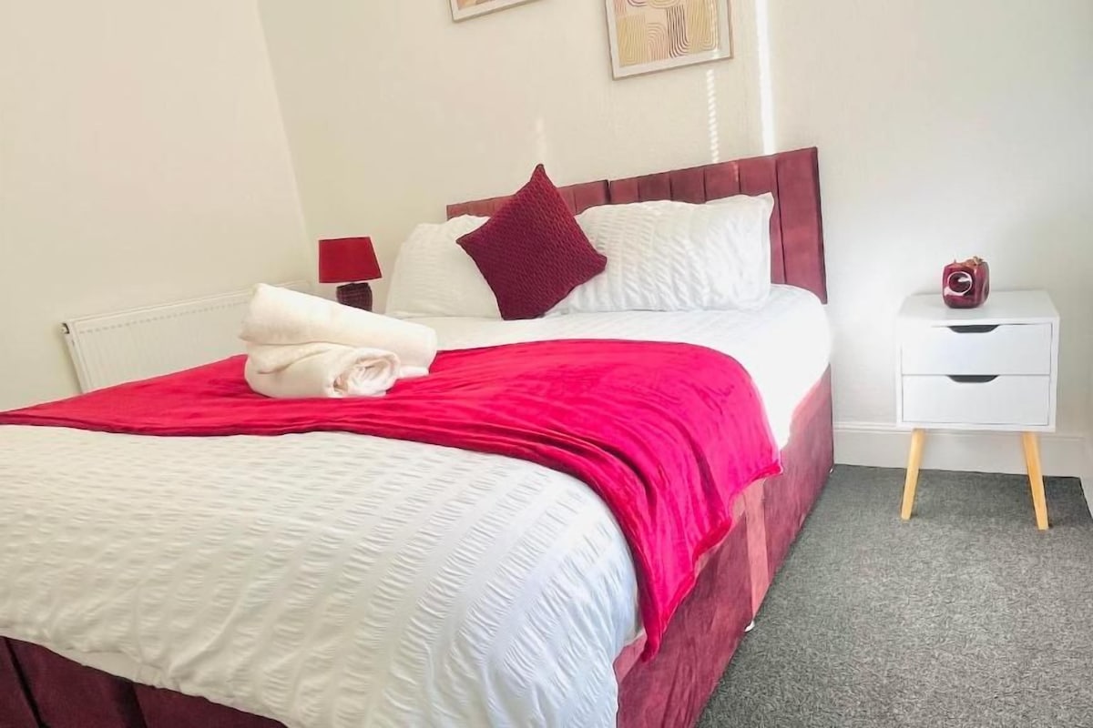 Een netjes opgemaakt bed met een rode hoofdbord, witte beddengoed en een rode plaid. Er staat een witte nachtkastje met een lamp en een klein decoratief object.
