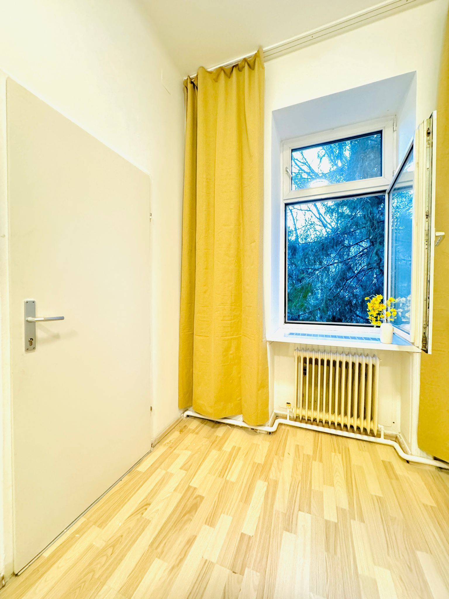 Een kamer met een houten vloer, een raam met uitzicht op bomen, een radiator en een geel gordijn.