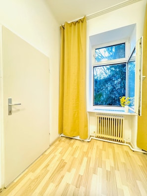 A room with a wooden floor, a window with a view of trees, a radiator, and a yellow curtain. - Your Vienna Key Private Room for Two (Wien)