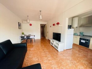 Una sala de estar moderna con un televisor de pantalla plana, un sofá negro, un comedor con sillas y una cocina con armarios y electrodomésticos.