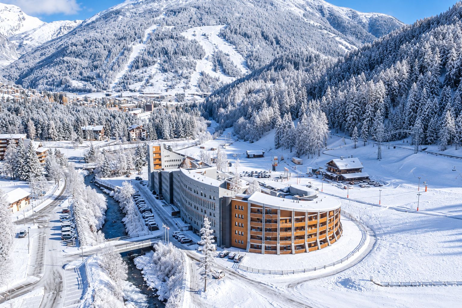 A snow-covered mountain town with a prominent round building with multiple floors and balconies, surrounded by snow-laden trees and a clear sky.