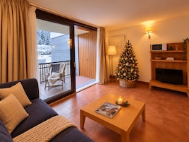 Una sala de estar con un sofá, una mesa de centro con una revista, un árbol de Navidad, un televisor sobre un mueble de madera y un ventanal corredizo que da a un área exterior.