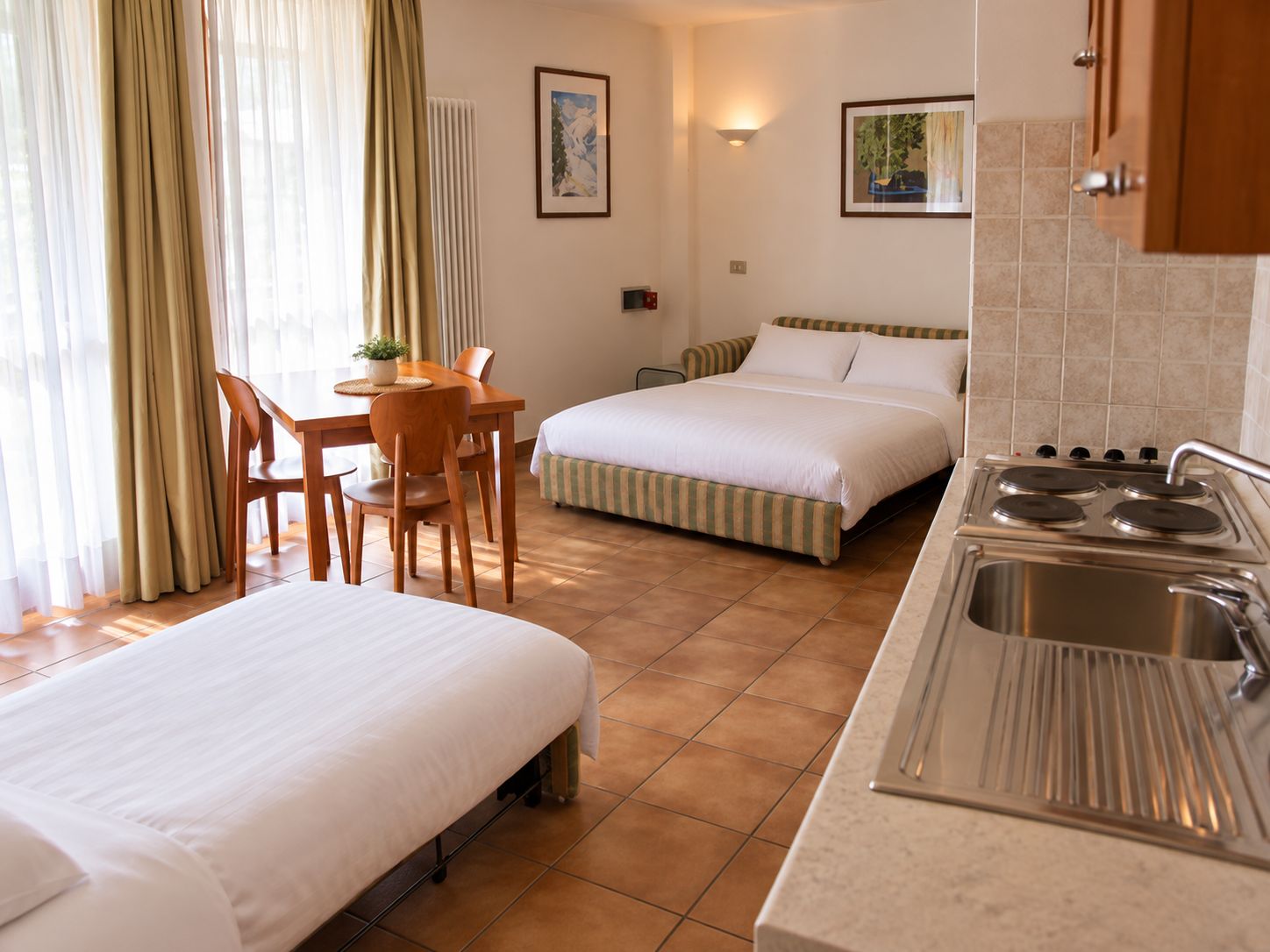 Habitación de hotel con dos camas, zona de comedor con mesa y sillas, kitchenette con estufa y fregadero, y ventana con cortinas.