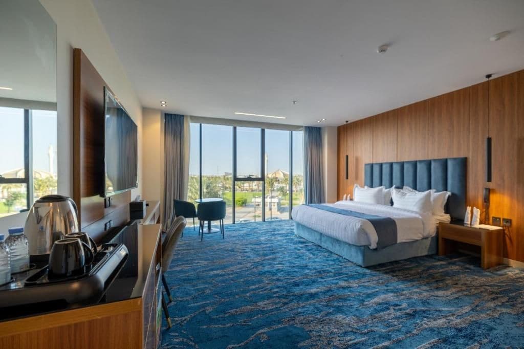 A hotel room with a large bed, a desk with a kettle, a chair, and a view of the outdoors.