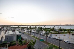 A coastal road with parked cars, palm trees, and a view of the sea and distant buildings. - Nasim Azraq Hotel (JEDDAH)
