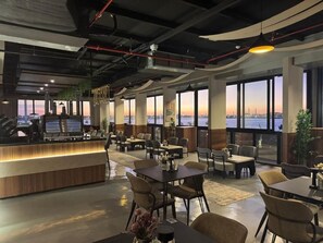A restaurant with a bar area, multiple tables, chairs, and large windows offering a view of the city skyline. - Nasim Azraq Hotel (JEDDAH)