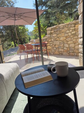 Ein Buch liegt auf einem Tisch, neben es steht eine Tasse. Ein Regenschirm und Sitzgelegenheiten im Freien sind zu sehen, im Hintergrund eine Steinmauer.