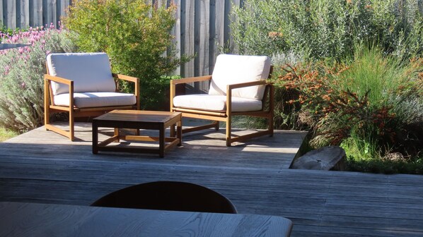 Un espacio al aire libre con dos sillas de madera y una mesita pequeña, rodeado de exuberante vegetación y una terraza de madera.