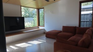 Una sala de estar con un televisor de pantalla plana grande montado en la pared, un sofá marrón y un ventanal con vistas a zonas verdes.