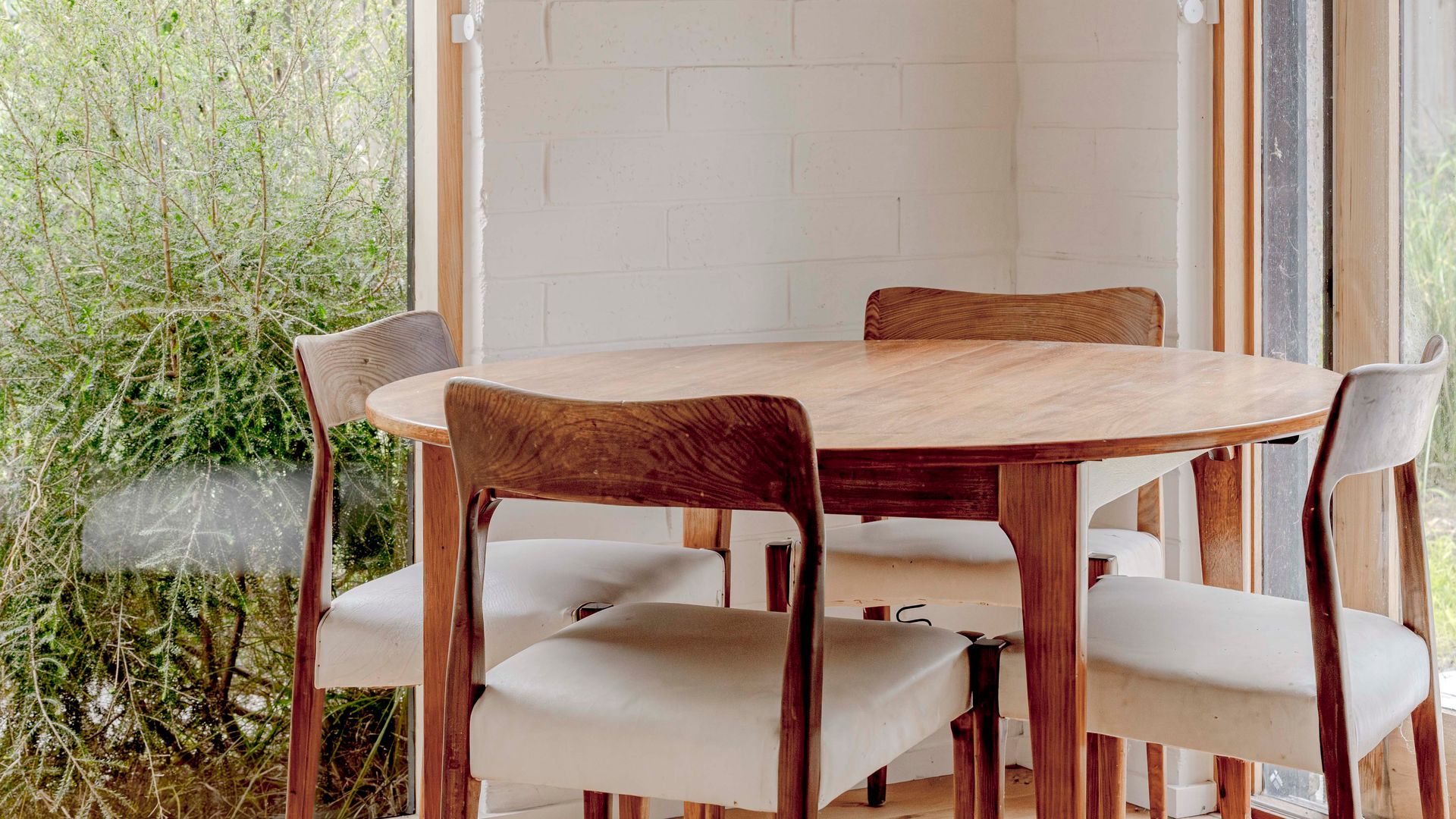 A round wooden dining table with six chairs, a white brick wall, and a view of greenery outside.
