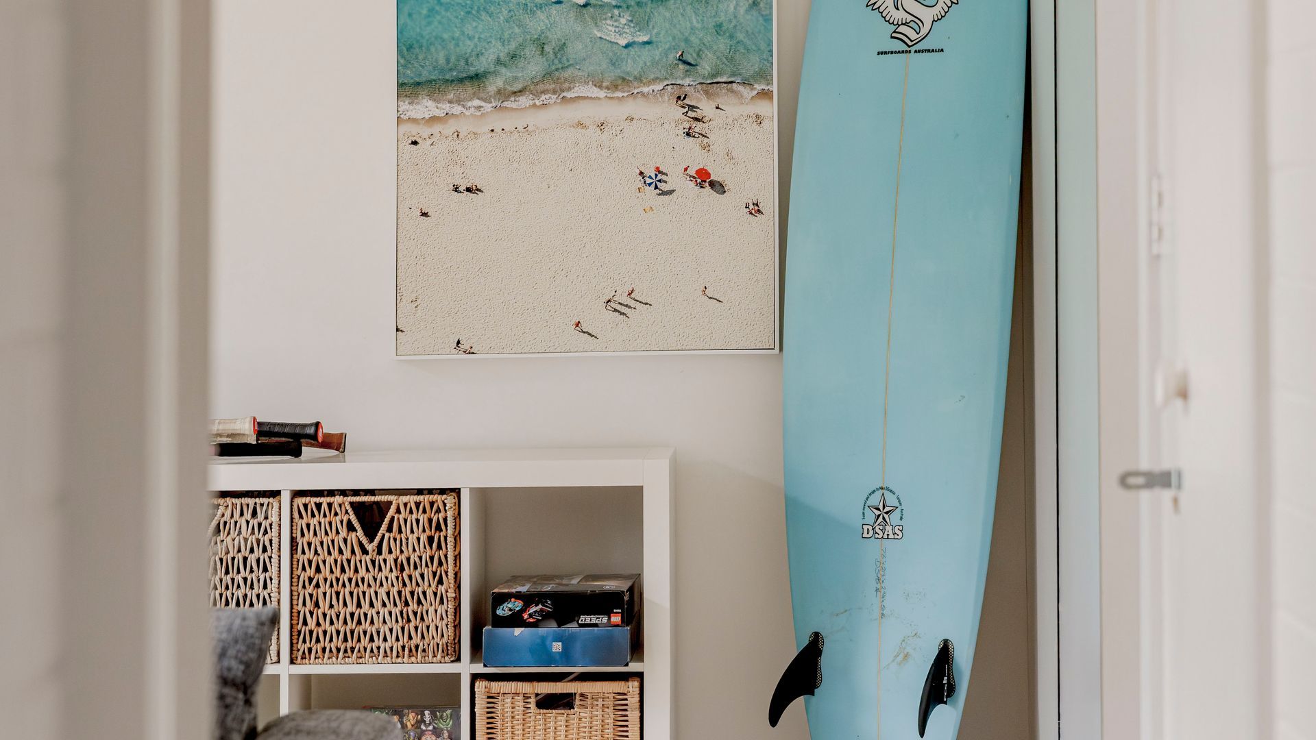 Una habitación con una tabla de surf apoyada en la pared, una fotografía enmarco de la playa y un estante blanco con diversos objetos.