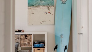 Una habitación con una tabla de surf apoyada en la pared, una fotografía enmarco de la playa y un estante blanco con diversos objetos.