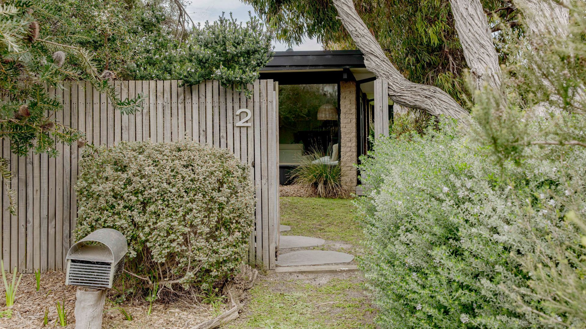 Una puerta de madera con el número 2, un buzón y un sendero que lleva a una zona exterior cubierta con un banco.