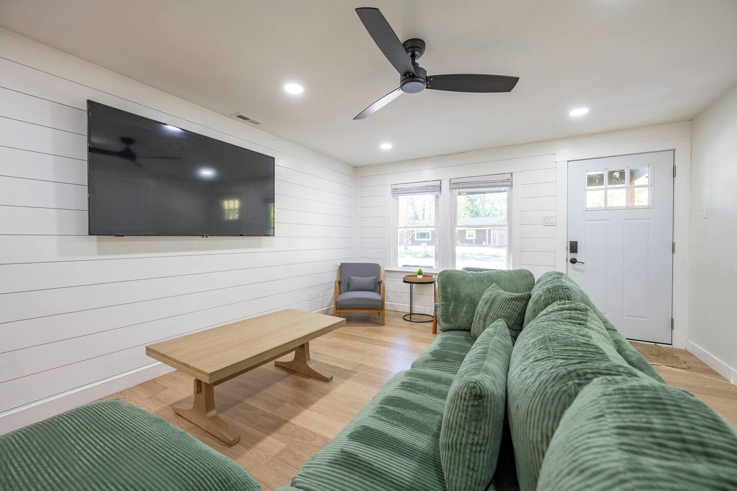 A living room with a flat-screen TV, a ceiling fan, a wooden table, a green sofa, and a door.