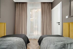 Hotellihuone, jossa on kaksi sänkyä, ovi, ikkuna, jossa on läpikuultavat verhot, ja valokytkin seinässä.