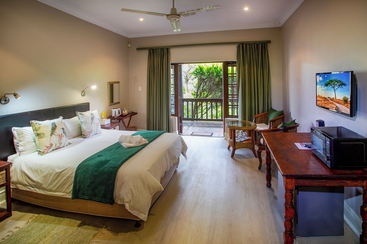 A hotel room with a bed, a desk, a television, and a balcony with a view of greenery.