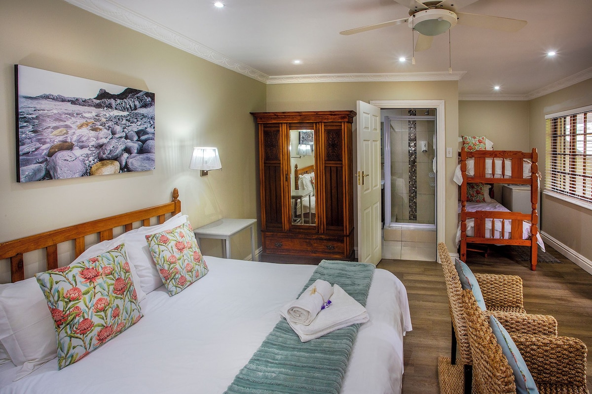 A hotel room with a bed, a mirror, a wooden wardrobe, a shower area, and a window with blinds.
