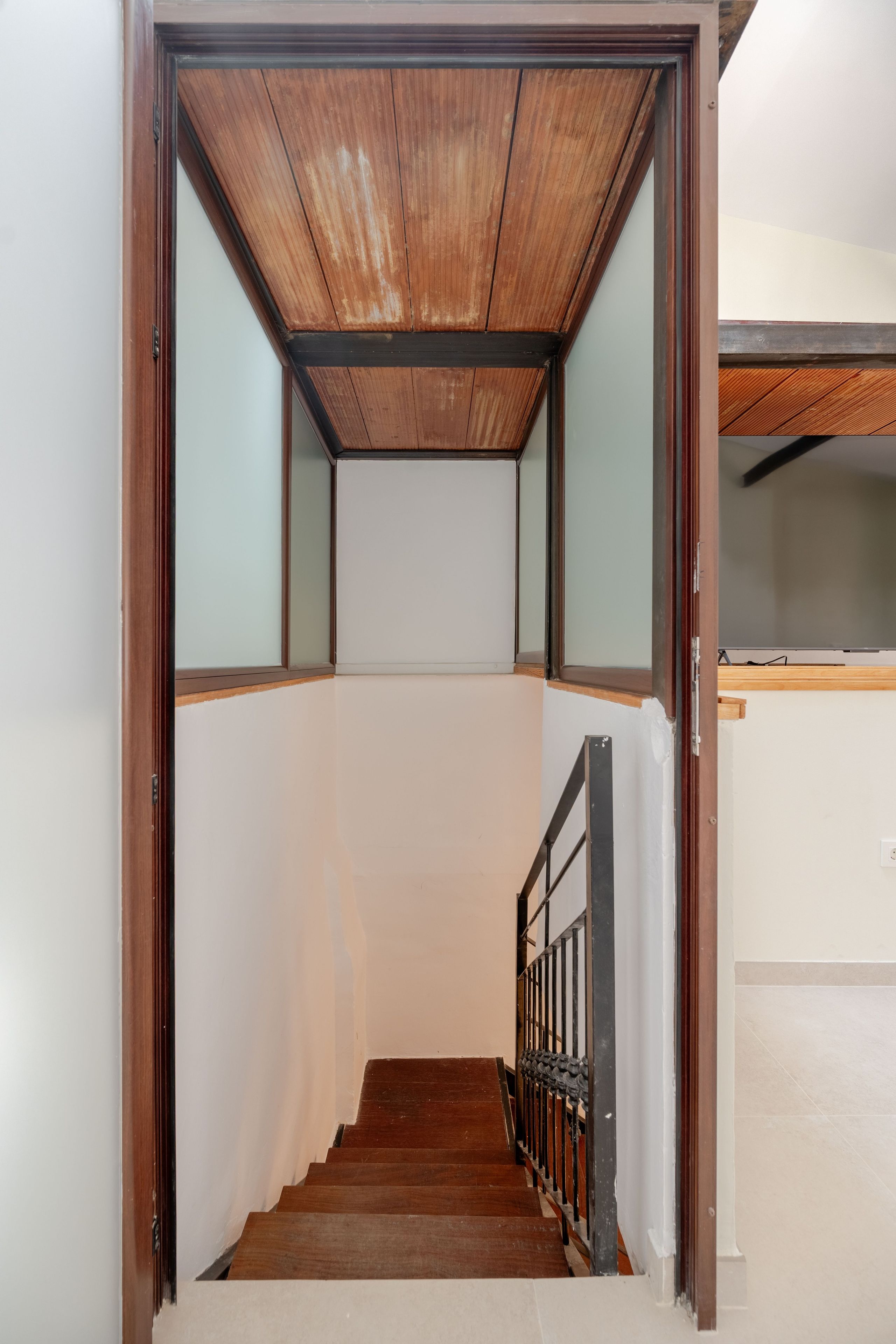 A staircase with wooden steps and a metal railing, leading to a room with a white wall and a wooden ceiling.
