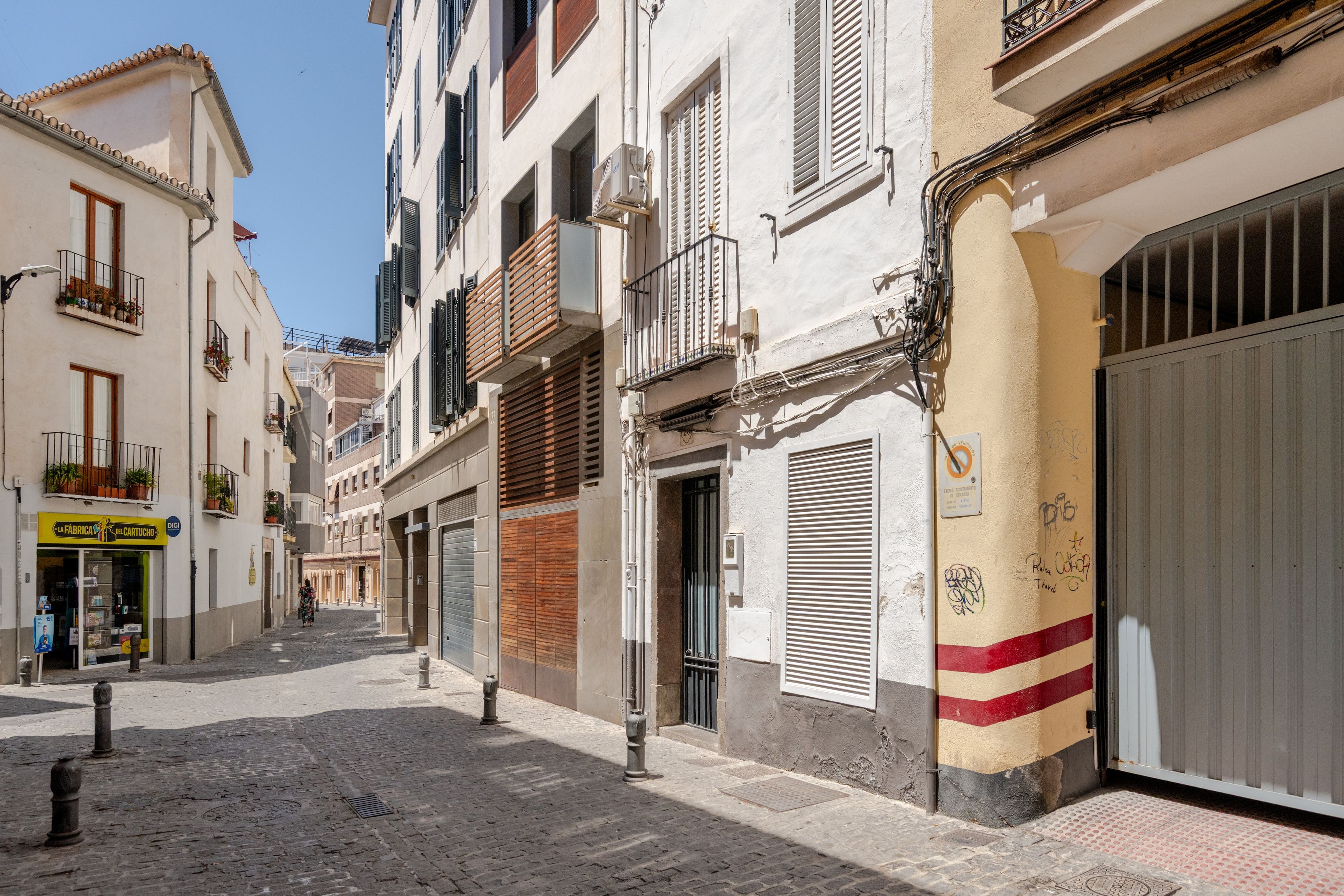 Una estrecha calle adoquinada flanqueada por edificios de varios pisos, algunos con balcones y contraventanas, y una tienda con un letrero.