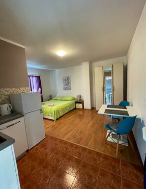 Une pièce compacte comprenant un coin cuisine, un lit avec une literie verte, une petite table avec une chaise et une porte donnant sur une autre pièce.