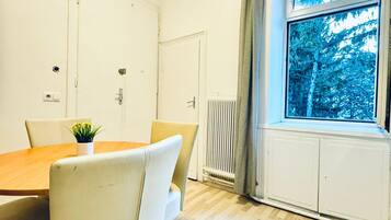 Ein Zimmer mit einem Holzesstisch, beigen Stühlen, einer Topfpflanze, einem Fenster mit Blick auf Bäume und einer Heizkörper.