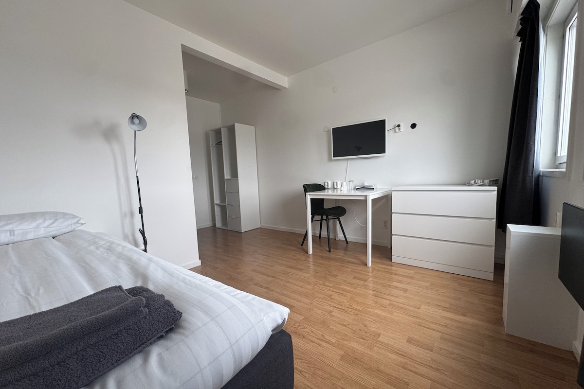 A modern hotel room with a bed, a desk, a chair, a flat-screen TV, and a wardrobe.