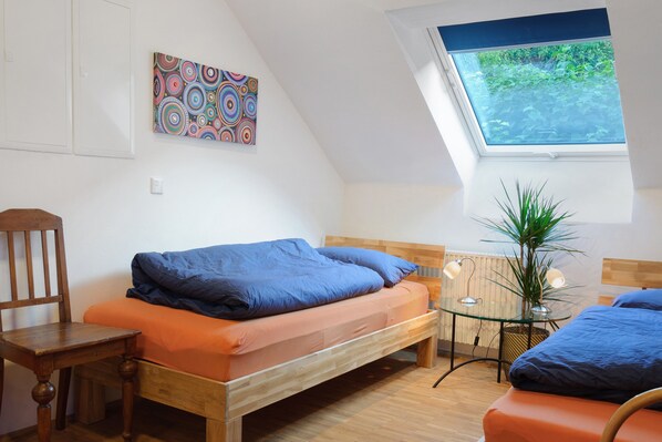 A bedroom with a bed, a chair, a small table, a potted plant, and a window with a view of greenery.