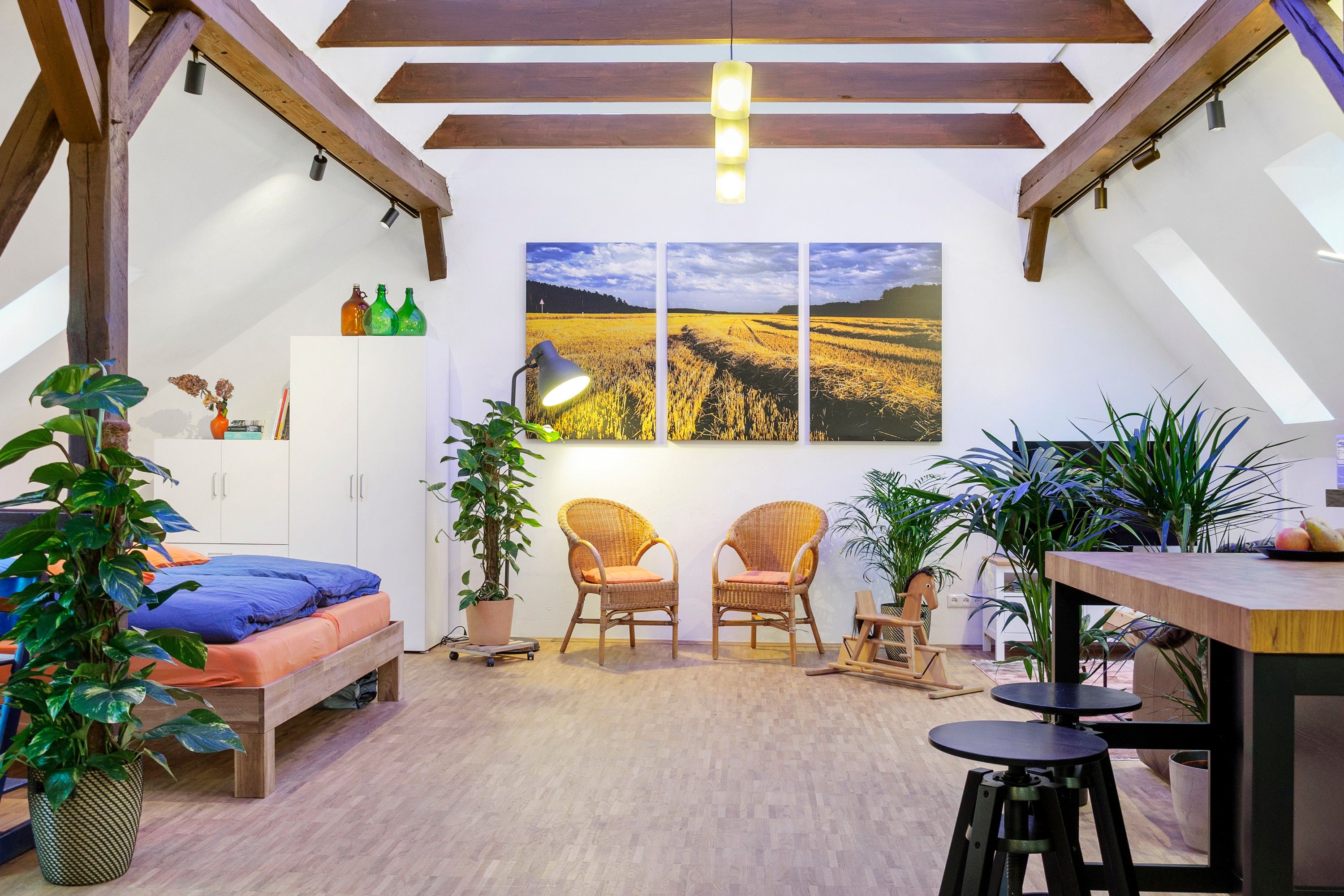 Une chambre à coucher aménagée dans un grenier, avec un plafond en pente, des poutres en bois, un lit, deux chaises, une table et une grande fenêtre ornée de trois tableaux représentant des paysages.