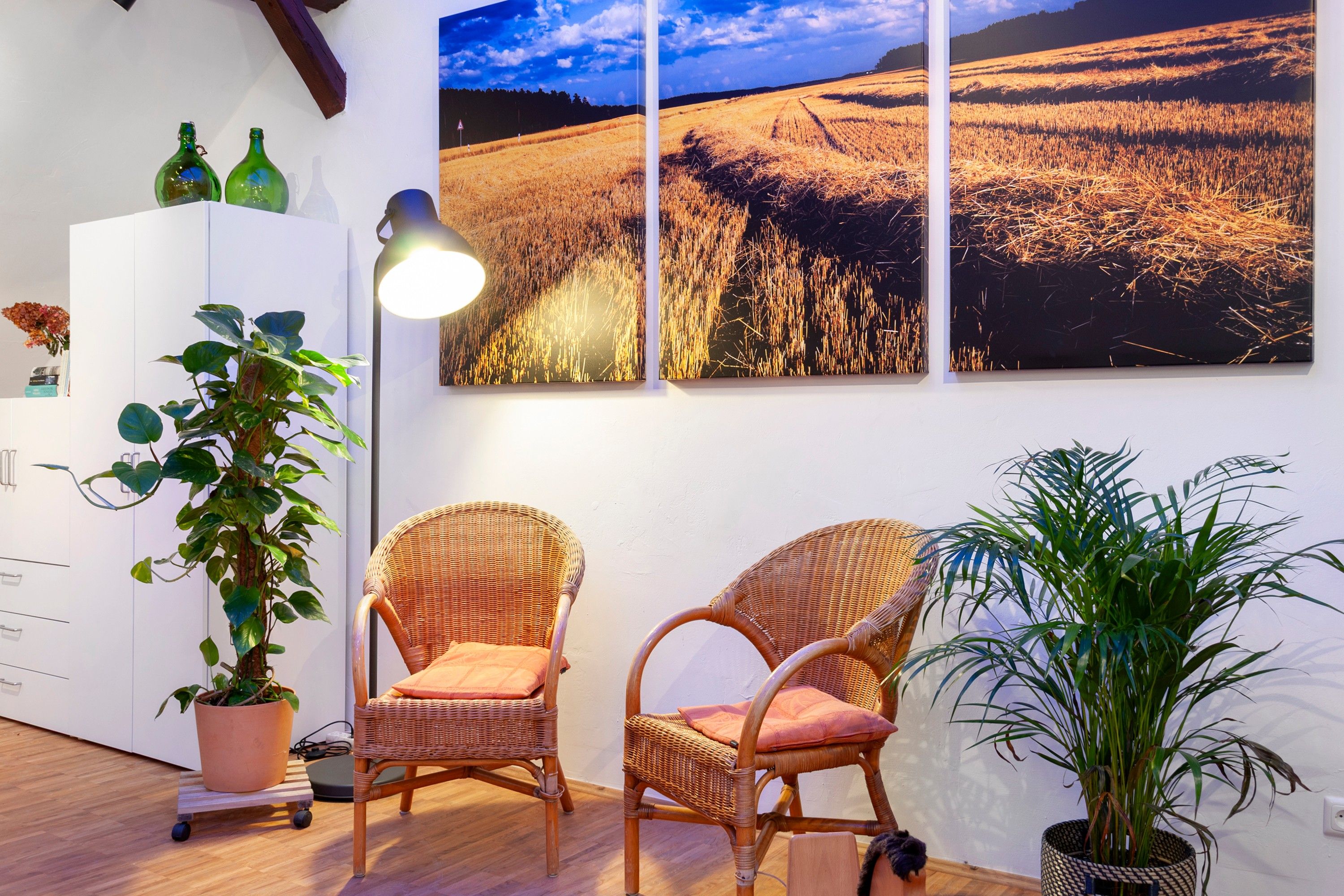 Ein Zimmer mit zwei Korbstühlen, einem Blumentopf, einem Schrank und einem großen Landschaftsbild an der Wand.