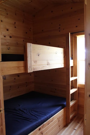 Ein Stockbett mit einer unterer Liege und einer Matratze, Holzwänden und einem Fenster.