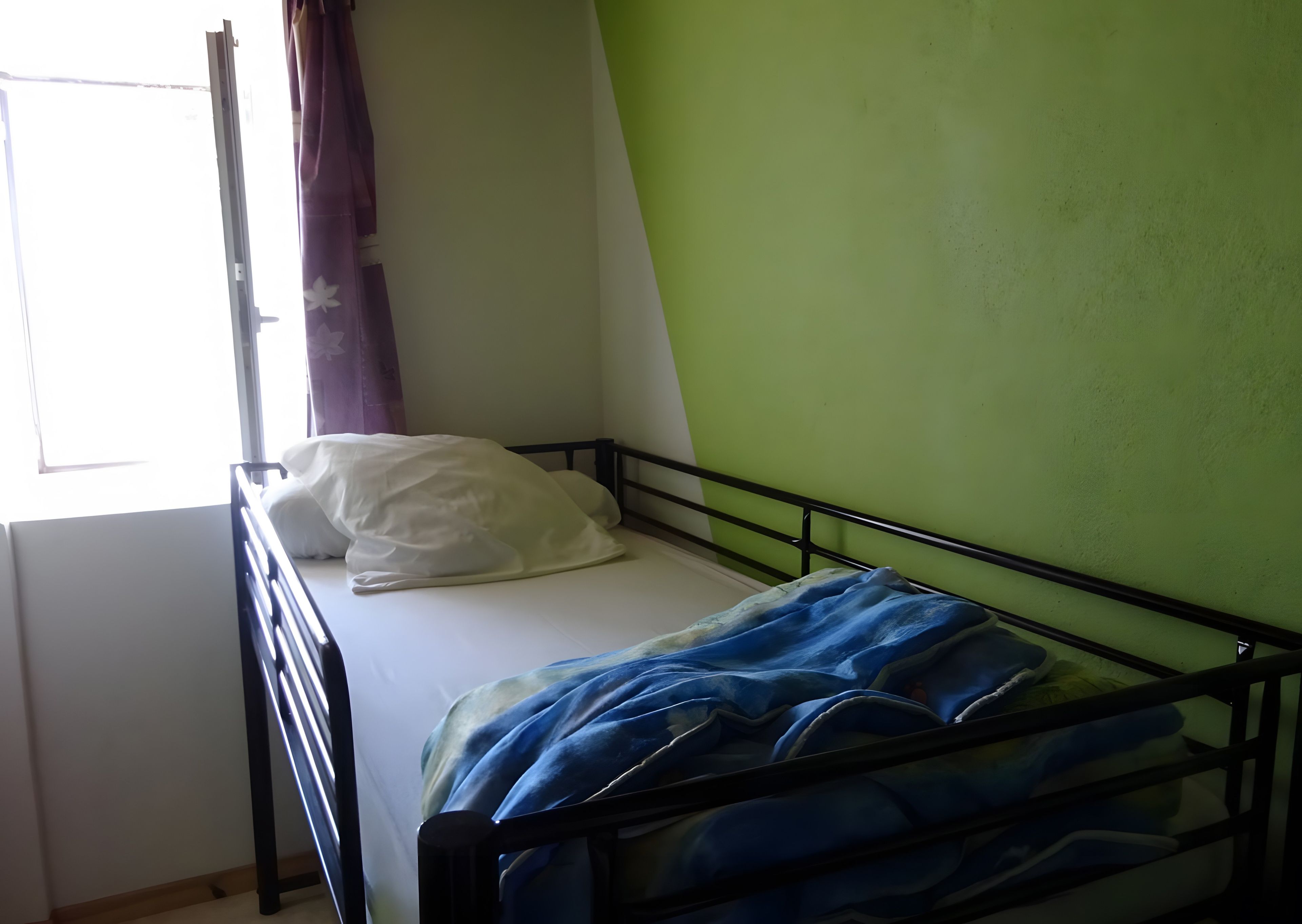 A single bed with a white pillow and a blue blanket, a window with curtains, and a green wall.